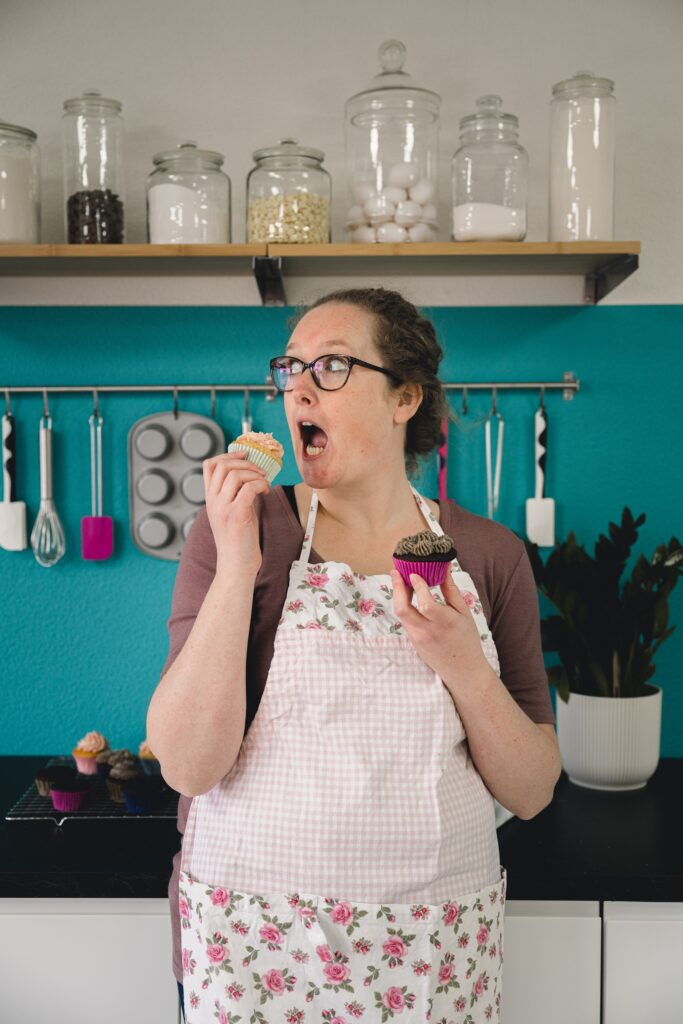 Roos in haar workshopruimte in Kampen, leunend tegen het aanrecht. Heeft een chocolade en een vanille cupcake vast. Ze kijkt opzij en doet alsof ze een cupcake gaat opeten.