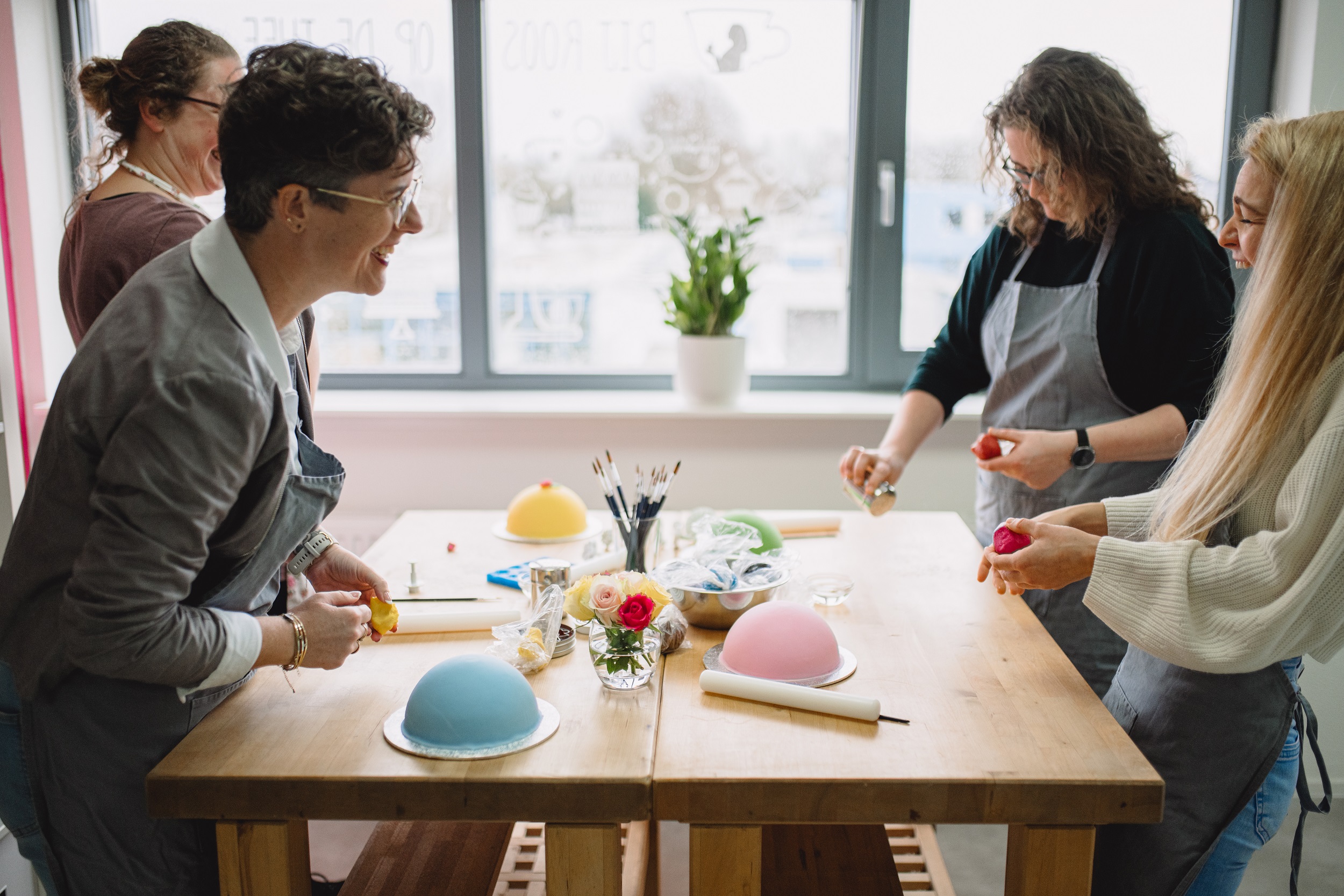 4 vrouwen met schorten die lachen terwijl ze boltaartjes aan het decoreren zijn tijdens een workshop